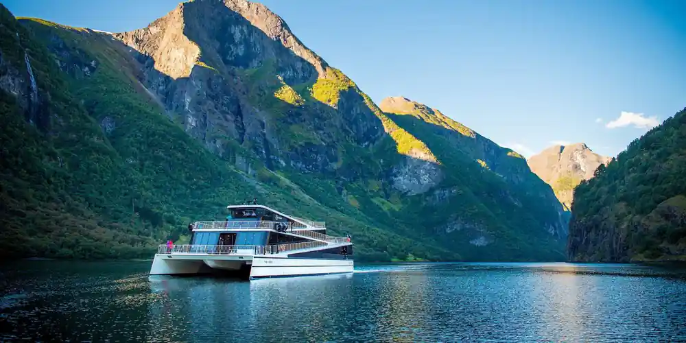 A modern electric fjord boat at the quay below a sunlit mountain